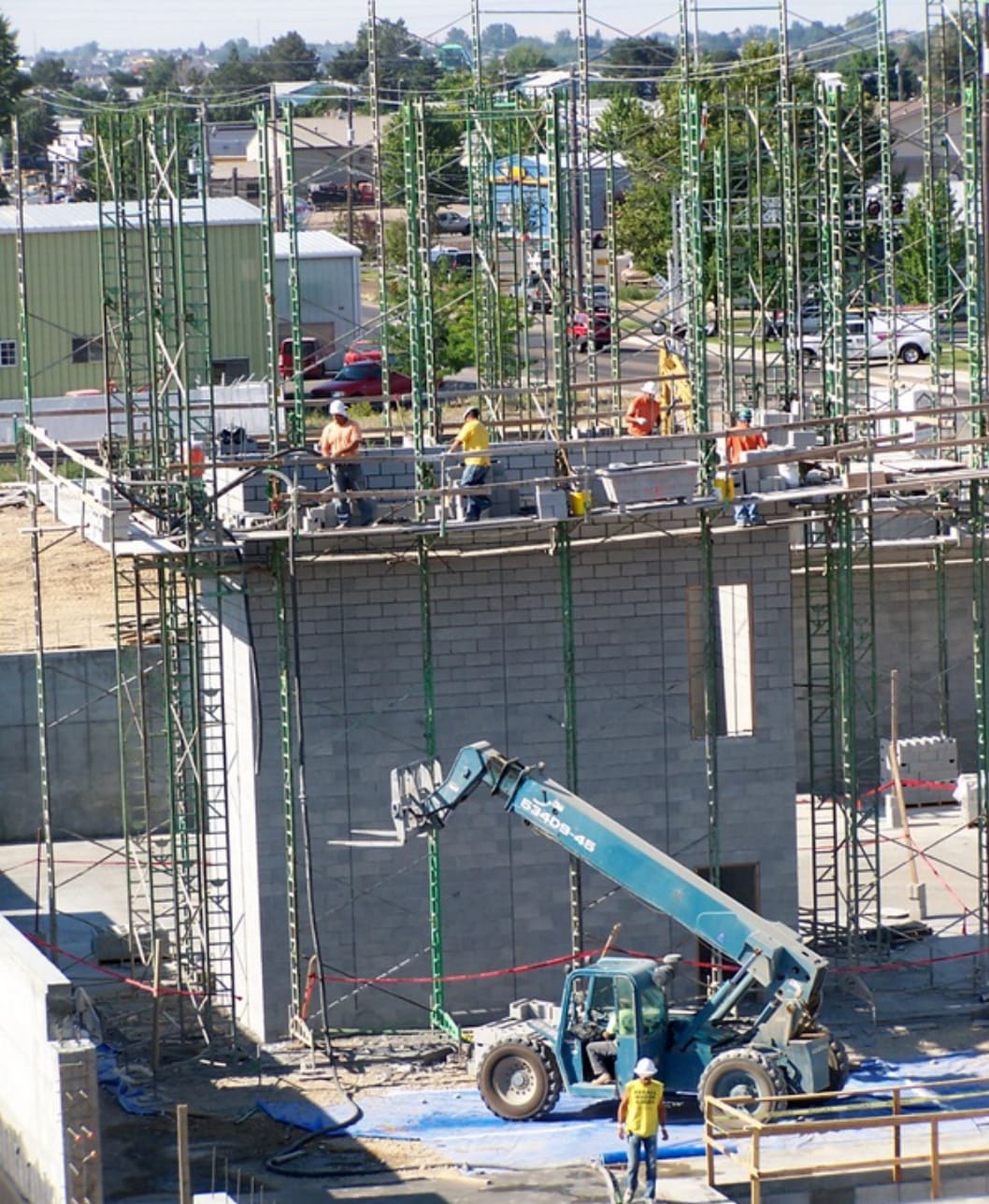 Masons laying concrete block walls on scaffolding at a commercial building site, with a telehandler below