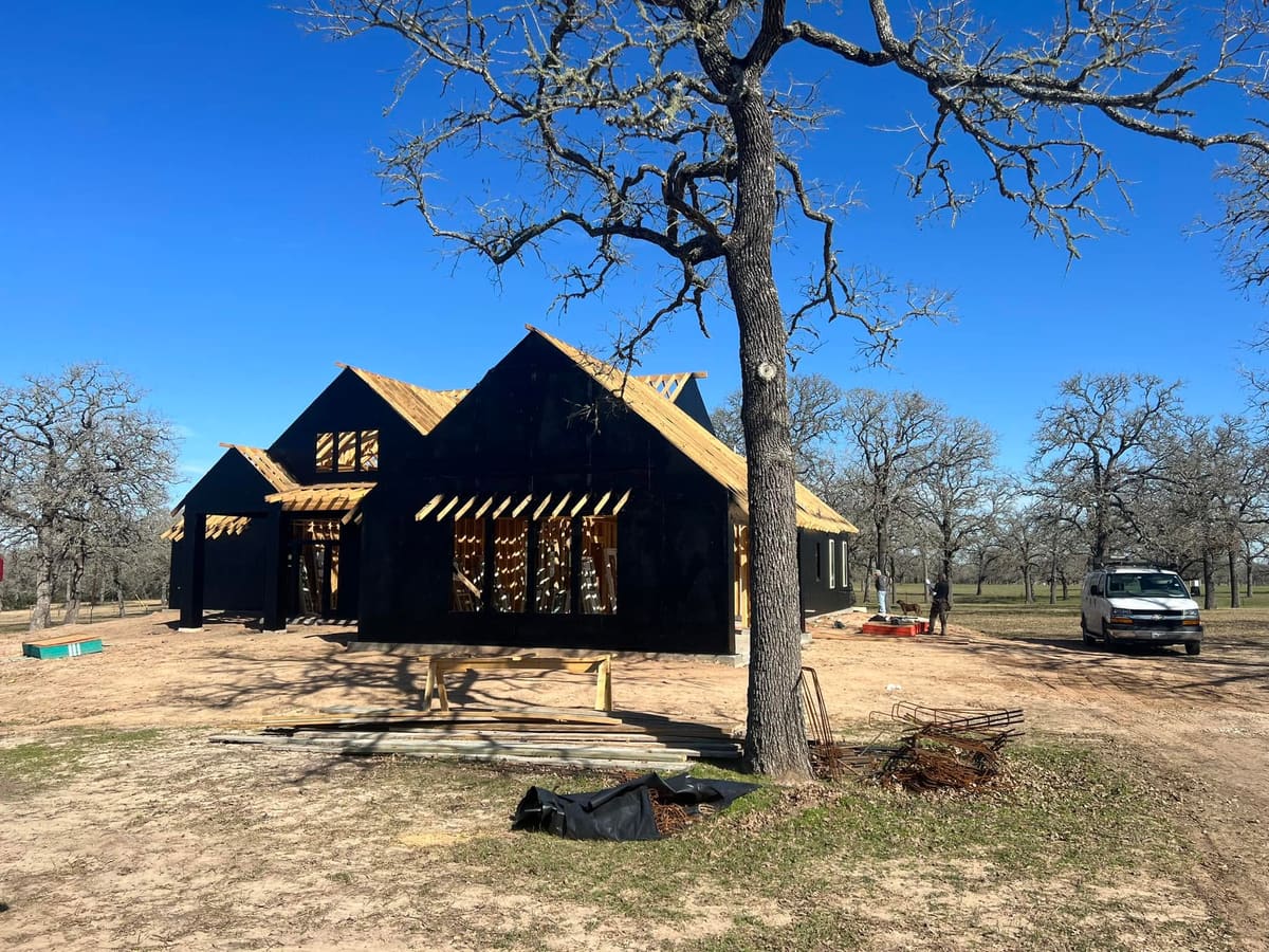 Two-story home wrapped in black weather barrier under a large oak tree