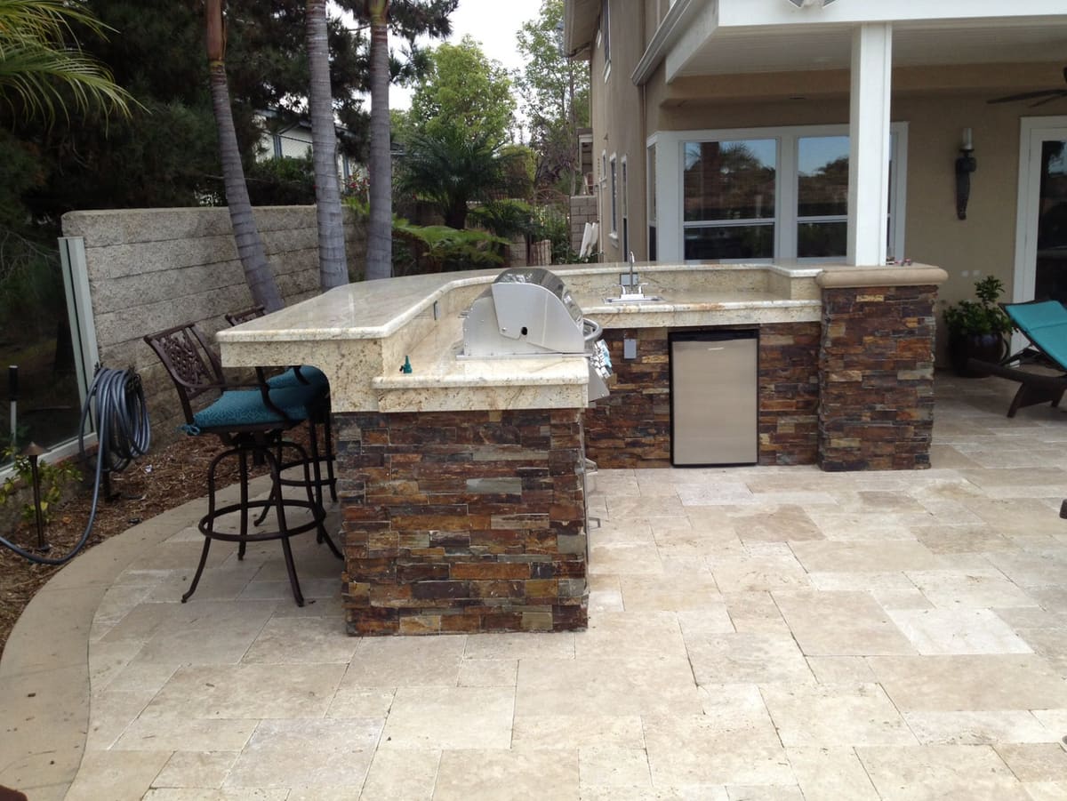Outdoor kitchen with a stone-clad bar, granite counters, a grill and a travertine patio